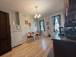 A kitchen with wooden cabinets, a refrigerator, a dining table with chairs, and a ceiling light fixture. - Cozy 1-bedroom apartment in charming Boston with high ceilings and bay windows (Boston)