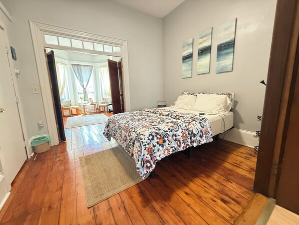 A bedroom with a wooden floor, a bed with a floral bedspread, a window with curtains, and a door leading to another room. - Cozy 1-bedroom apartment in charming Boston with high ceilings and bay windows (Boston)