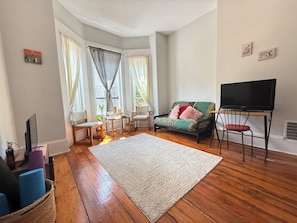 A living room with a sofa, TV, and wooden flooring. - Cozy 1-bedroom apartment in charming Boston with high ceilings and bay windows (Boston)