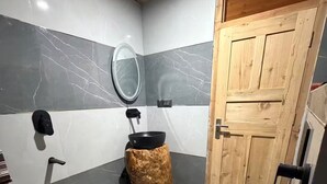 Un baño con puerta de madera, un inodoro, un lavabo con espejo y un lavatambos negro sobre un taburete.