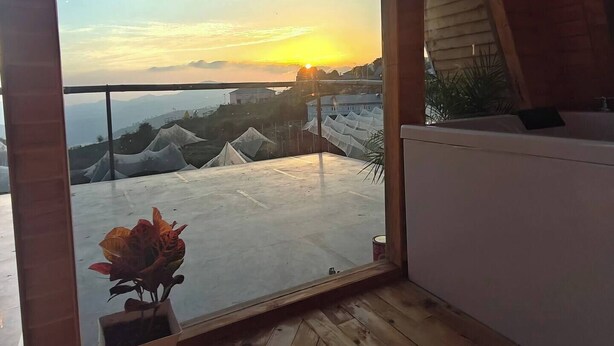 Una habitación con un amplio ventanal que da a una vista panorámica al atardecer, suelo de madera y una planta en maceta.