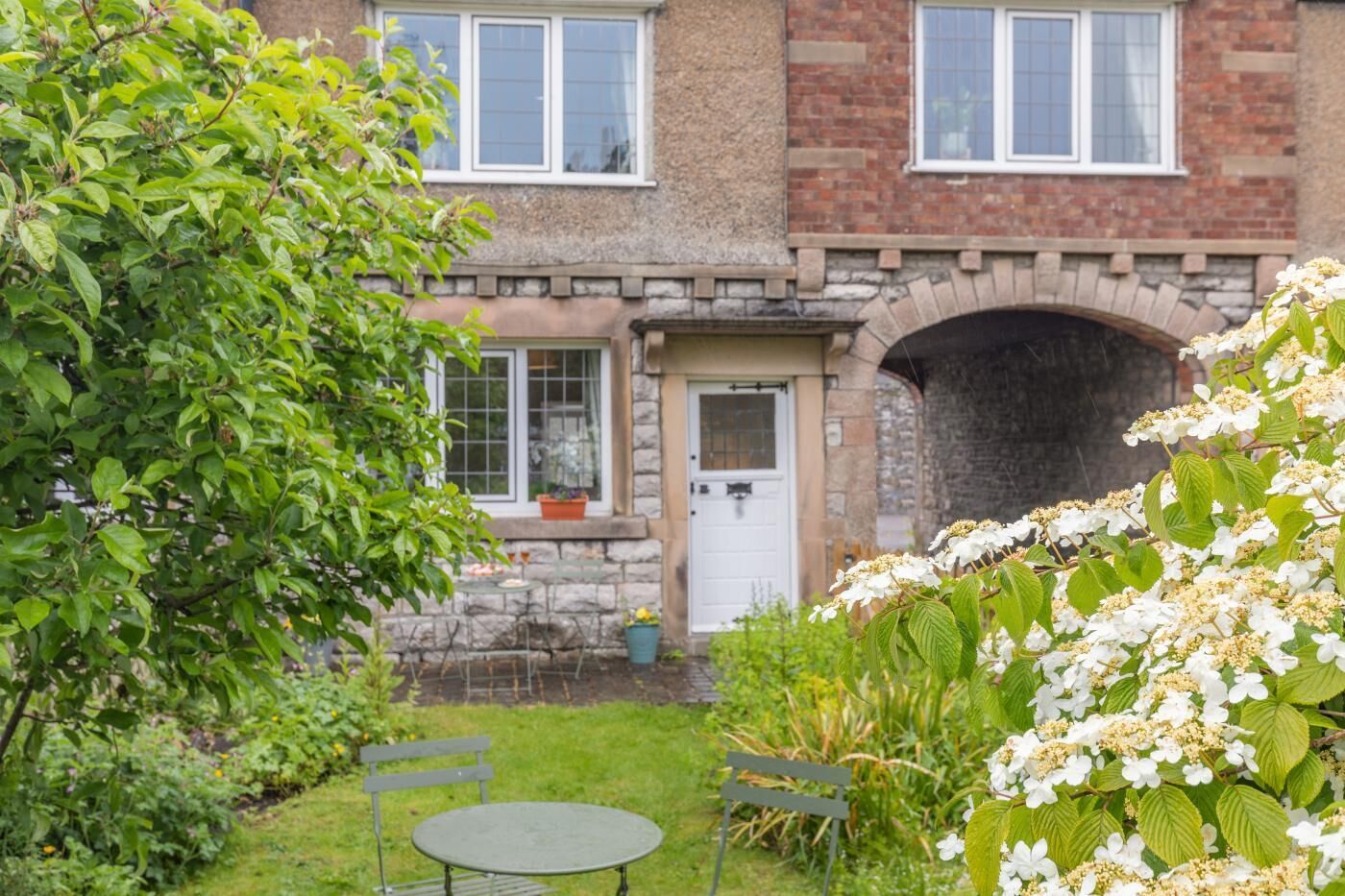 Une maison en briques avec un portail en pierre, une porte d’entrée et un petit jardin agrémenté d’un banc et d’une table.