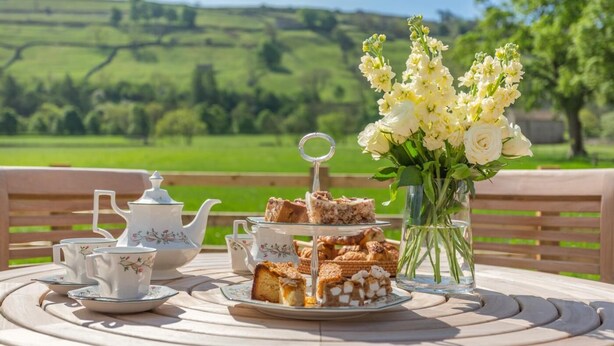 A table set with a teapot, cups, a tray of pastries, and a vase of flowers, with a scenic view in the background. - 1 Bed in Low Row (oc-c43317) (Low Row)
