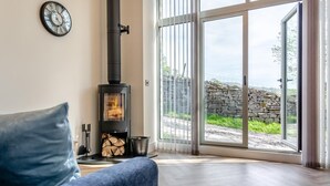 A living room with a modern fireplace, a clock on the wall, and a view of a stone wall outside. - 1 Bed in Low Row (oc-c43317) (Low Row)