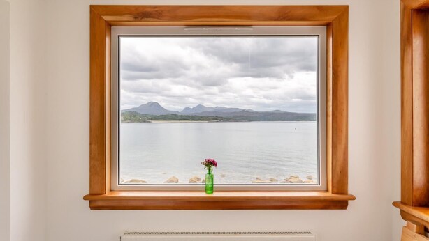A window with a view of a lake and mountains, a radiator below, and a green vase with flowers on the sill. - 2 Bed in Strath (oc-s44223) (Gairloch)
