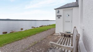 A white building with a door, a bench, and a view of a calm body of water. - 2 Bed in Strath (oc-s44223) (Gairloch)