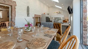Un coin repas avec une table en bois dressée pour le repas, une cuisine en arrière-plan et un mur en pierre.