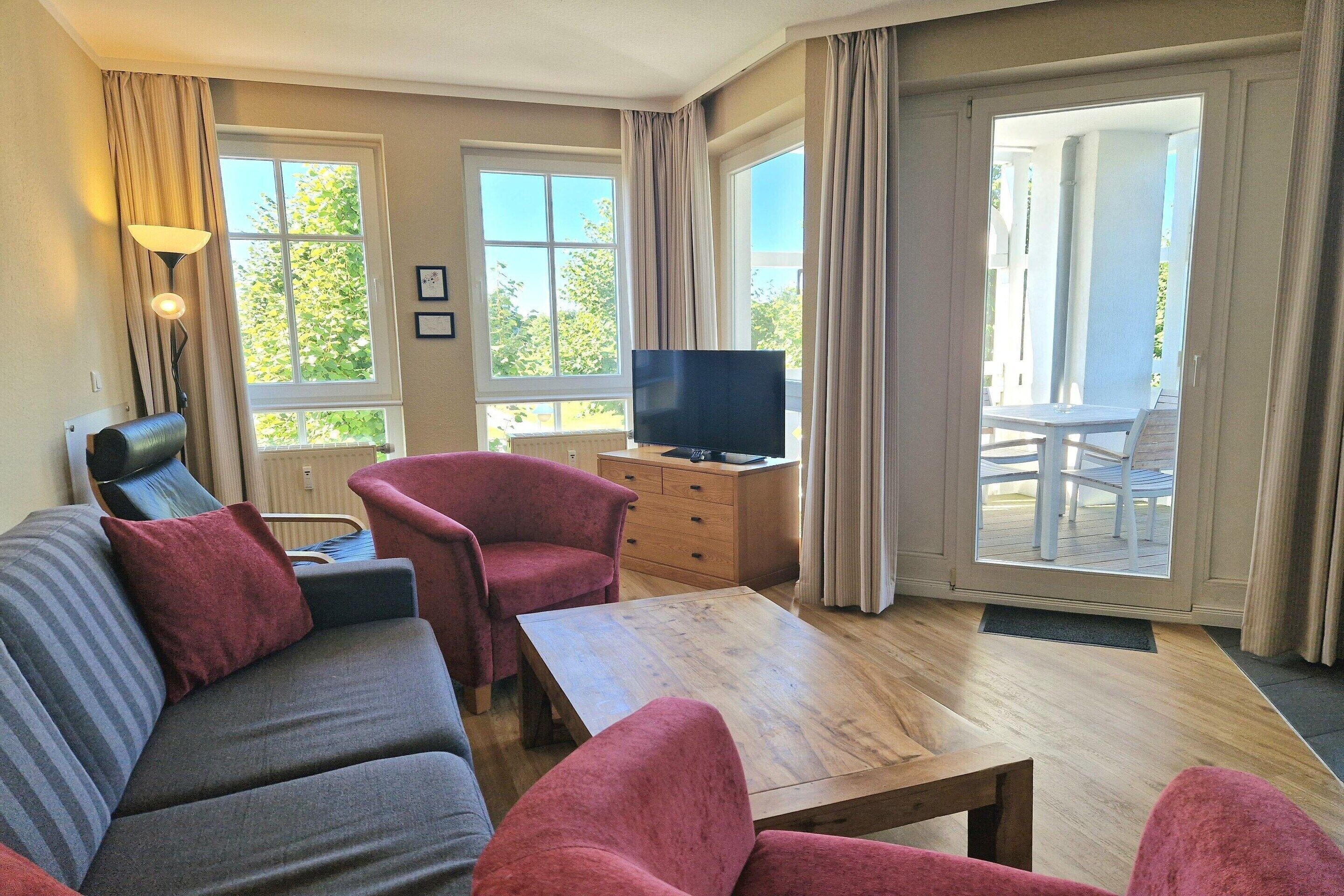 Ein Wohnzimmer mit Sofa, Sesseln, Couchtisch, Fernseher und Blick ins Freie.