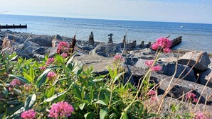 Eine felsige Küstenlinie mit freiem Blick auf das Meer, eine kleine Struktur mit einem Kreuz und rosa Blüten im Vordergrund.