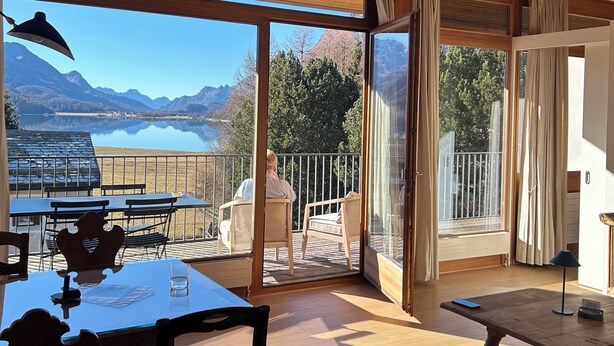 Ein Holzinnenraum mit Tisch und Stühlen, Blick auf Berge und See durch große Fenster, und ein Balkon mit Geländer.