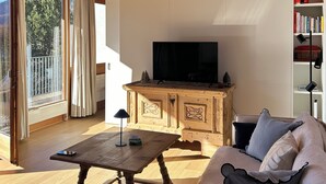 Eine Holzbalkendecke, ein Fernseher auf einem Schrank, ein Sofa, ein Tisch und ein Bücherregal.