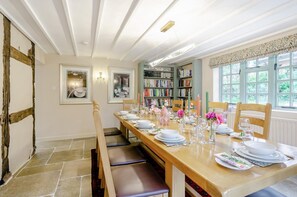 A dining room with a long wooden table set for six, flanked by bookshelves, framed pictures, and a window with a view of greenery. - 4 Bed in Radford (oc-r33951) (Rous Lench)