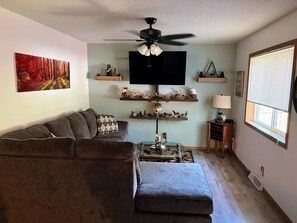 A living room with a sectional sofa, a glass coffee table, a ceiling fan, a flat-screen TV, decorative shelves, and a window with blinds. - Secluded Woodland Retreat for 6 Near Wisconsin Dells, Lakes & ATV Trails (New Lisbon)