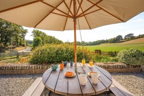 A round wooden table with a beige umbrella, surrounded by a stone wall, with a view of a green field and trees in the background. - 3 Bed in Tywyn (oc-e43276) (Tywyn)