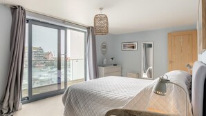 Une chambre à coucher comprenant un lit, une commode, un miroir, un luminaire suspendu et une vue sur un port de plaisance, visible à travers une grande fenêtre.