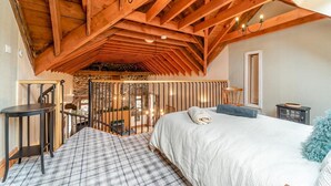 Una camera da letto con soffitto alto, travi in legno, un letto con copripiumino bianco, un tavolino e vista su un muro in pietra e un balcone.