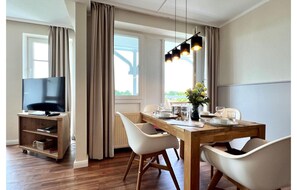 A dining area with a wooden table, white chairs, a television on a stand, and a window with curtains. - Urlaub mit Haustier in Sellin (Sellin)