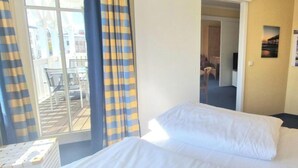 A hotel room with a bed, a window with curtains, a view of the outside, and a door leading to another room. - Urlaub auf Rugen mit Hund (Sellin)