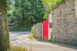 A stone wall with a red gate, a paved road, and dense greenery in the background. - 2 Bed in Yealmpton (oc-o42800) (Yealmpton)
