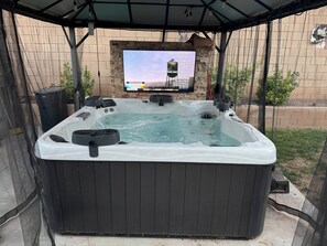 Un jacuzzi con una pantalla de televisión que muestra un noticiero, rodeado por una zona cubierta con pared de ladrillo y follaje.