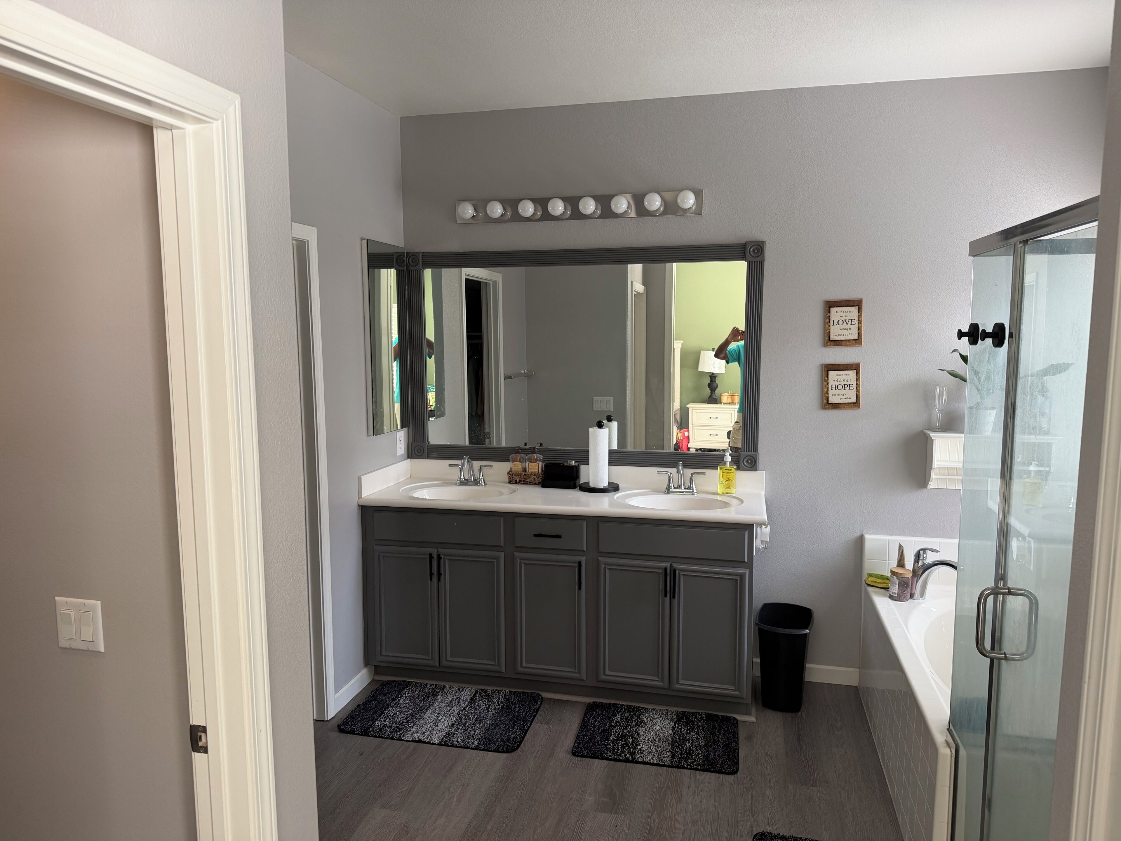 A bathroom with a double vanity, a large mirror, a glass shower enclosure, and a black trash bin.