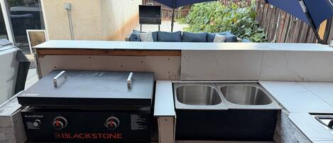 Cocina exterior con una parrilla Blackstone, un lavamanos y una zona de estar bajo un paraguas azul.