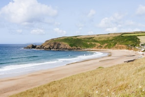 Paisagem costeira com uma praia arenosa, áreas gramadas e um afloramento rochoso.