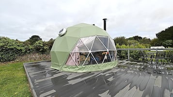 Una tienda en forma de domo geodésico con chimenea, ubicada sobre una terraza de madera y un muro de piedra, con vegetación de fondo.