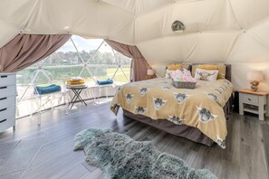 A tented room with a bed, a small table, a chair, and a view of the outdoors.