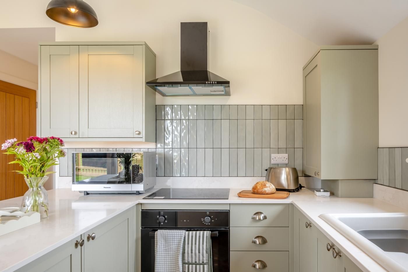 Una cocina moderna con horno empotrado, microondas y tostadora, con armarios de colores claros y una campana extractora de acero inoxidable.