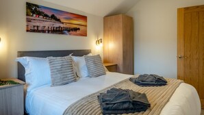 A neatly made bed with pillows and a blanket, a wooden wardrobe, a wooden door, and a wall-mounted picture of a sunset over water. - 2 Bed in Adgestone (oc-i42801) (Brading)