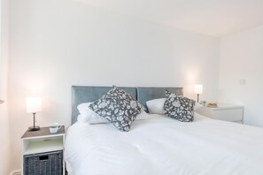 A neatly made bed with a grey headboard, two bedside lamps, a small bedside table with a cup and a plate of food, and a wicker basket under the nightstand. - 2 Bed in Rhoslefain (oc-u44707) (Rhoslefain)