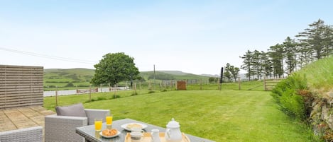 An outdoor patio with wicker furniture, a table set with breakfast items, and a view of a grassy field and trees.