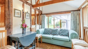 Um quarto aconchegante com vigas de madeira aparentes, um sofá azul claro, uma mesa com um vaso de flores e vista para o jardim através de janelas amplas.