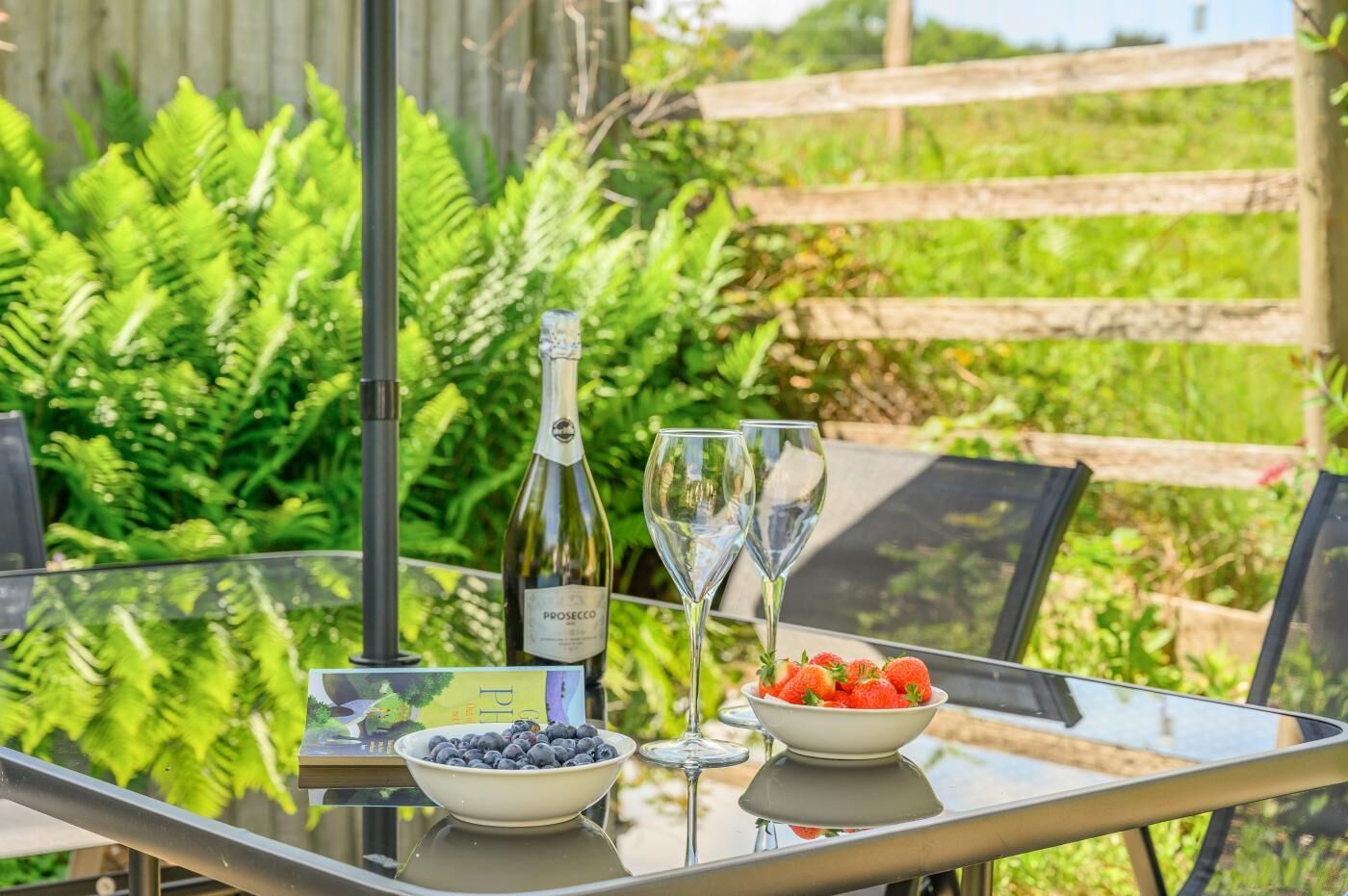 A glass table with a bottle of wine, two wine glasses, a bowl of strawberries, and a bowl of blueberries.