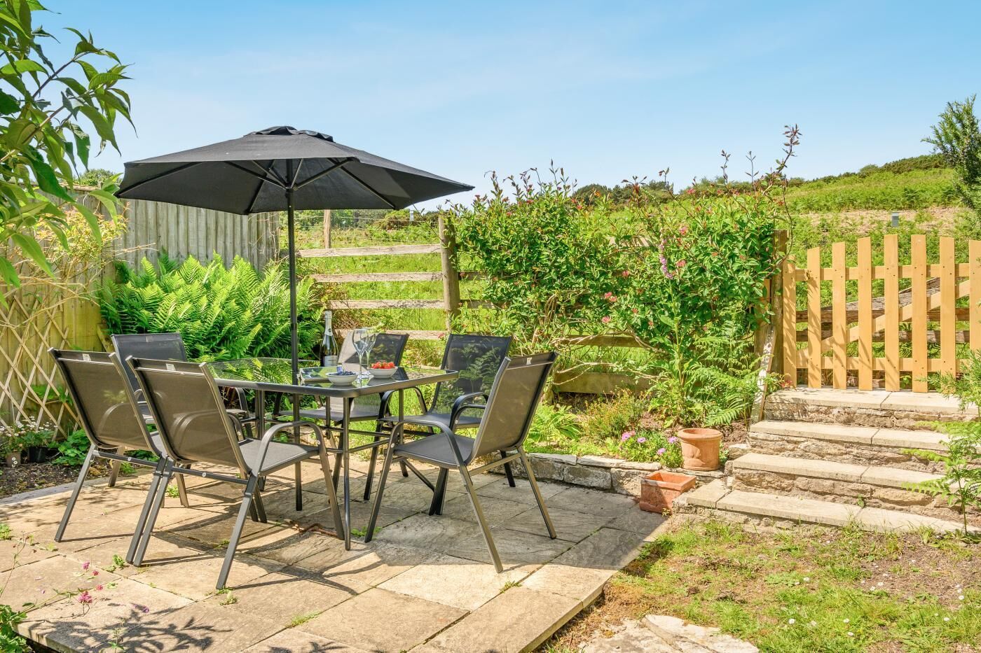 En udendørs terrasse med et bord og stole, en sort parasol og et træhegn.