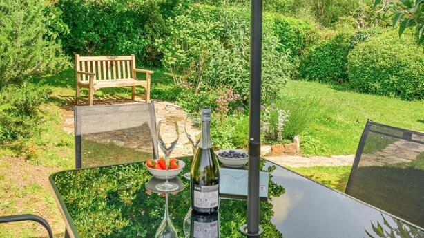 A patio with a table, a bottle, a bowl of strawberries, and a bench in a lush garden. - 3 Bed in Studland (oc-o42812) (Studland)