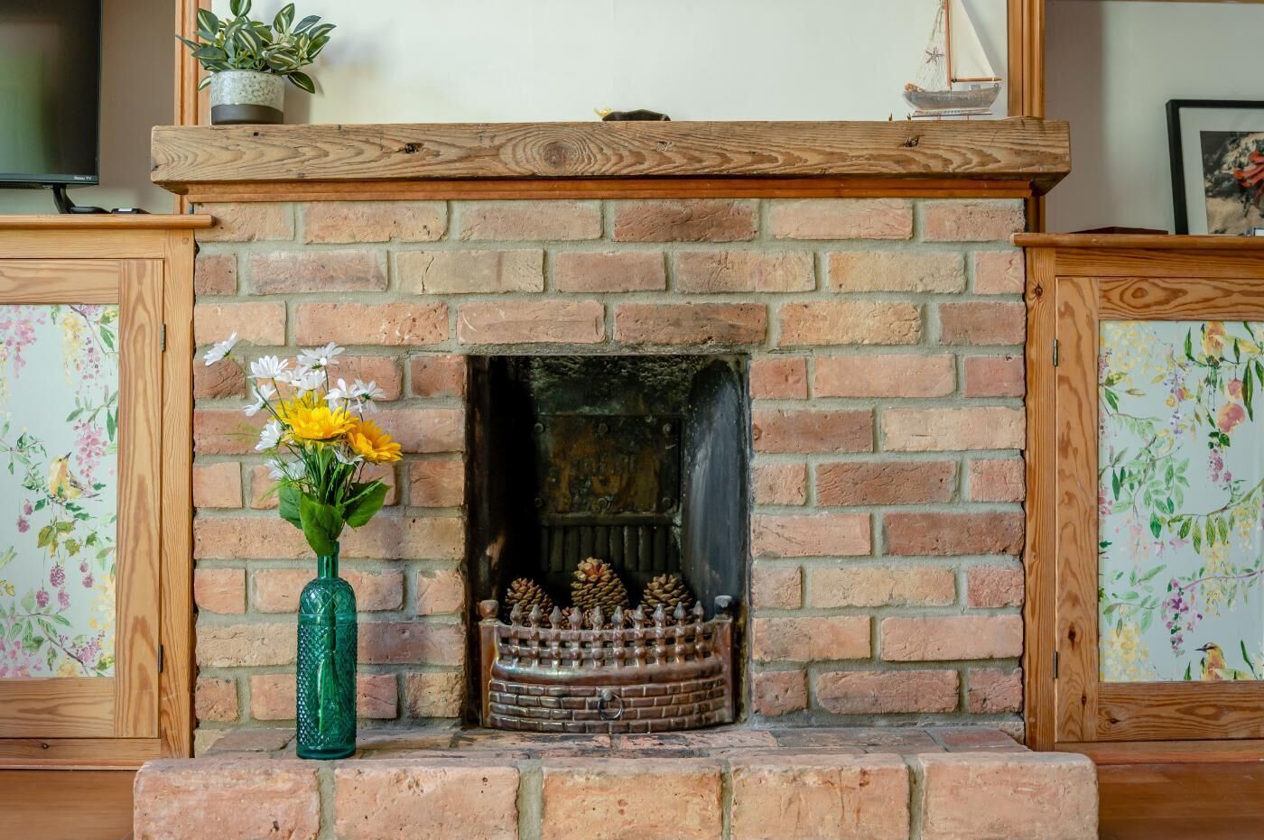 A brick fireplace with a wooden mantel, a green vase with flowers, and a birdcage with pinecones.