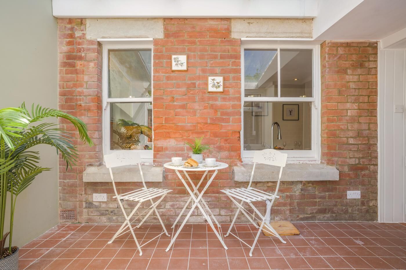 A patio with two white chairs and a small table, a potted plant, and a brick wall with windows.