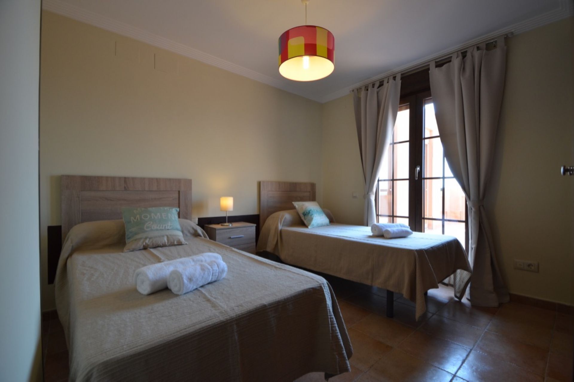 A hotel room with two beds, a nightstand, a lamp, and a window with curtains.