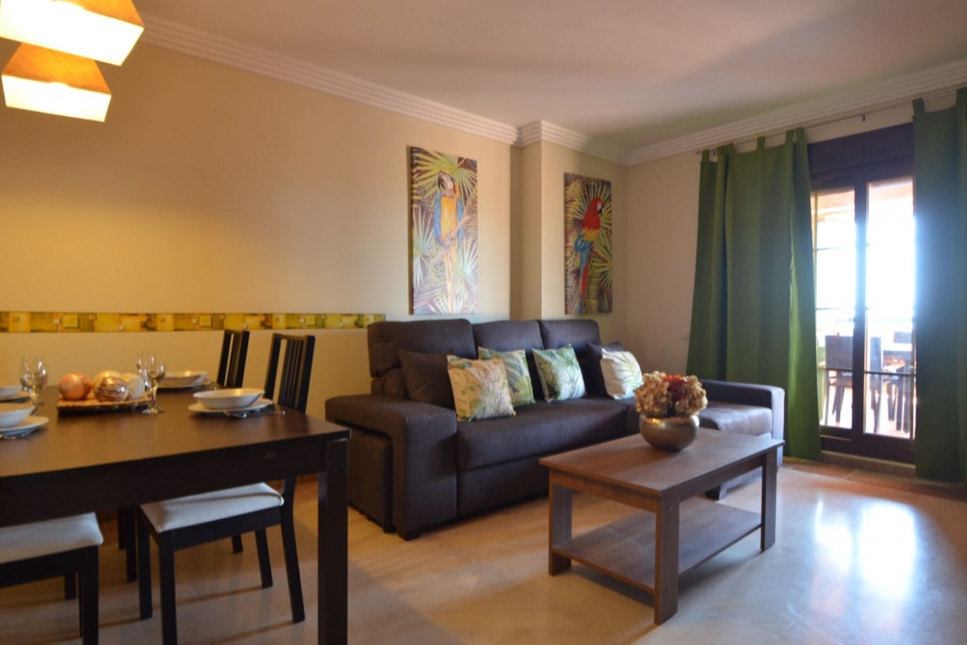 A living room with a dark sofa, a coffee table, and a dining table set with plates and glasses. There are two paintings on the wall and a window with green curtains.