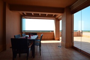 Ein Essbereich mit Tisch und Stühlen, Meerblick durch große Fenster und ein Balkon mit Geländer.