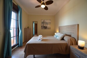 Ein Hotelzimmer mit Bett, Nachttisch, Deckenventilator, einem Fenster mit Vorhängen und einem Bild an der Wand.