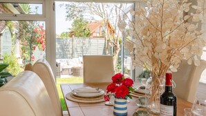 Un coin repas avec une table dressée pour deux personnes, un vase rempli de fleurs rouges, une bouteille de vin et une vue sur un jardin, visible à travers de grandes fenêtres.