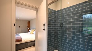 A modern bathroom with dark blue tiles, a glass shower enclosure, and a visible bedroom with a bed and a nightstand. - 2 Bed in Sibton (oc-q43760) (Sibton)