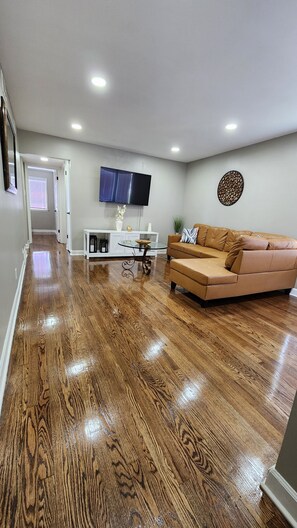 A spacious living room with hardwood flooring, a brown leather sofa, a glass coffee table, and a flat-screen TV mounted on the wall. - Stylish Midway Getaway (Chicago)