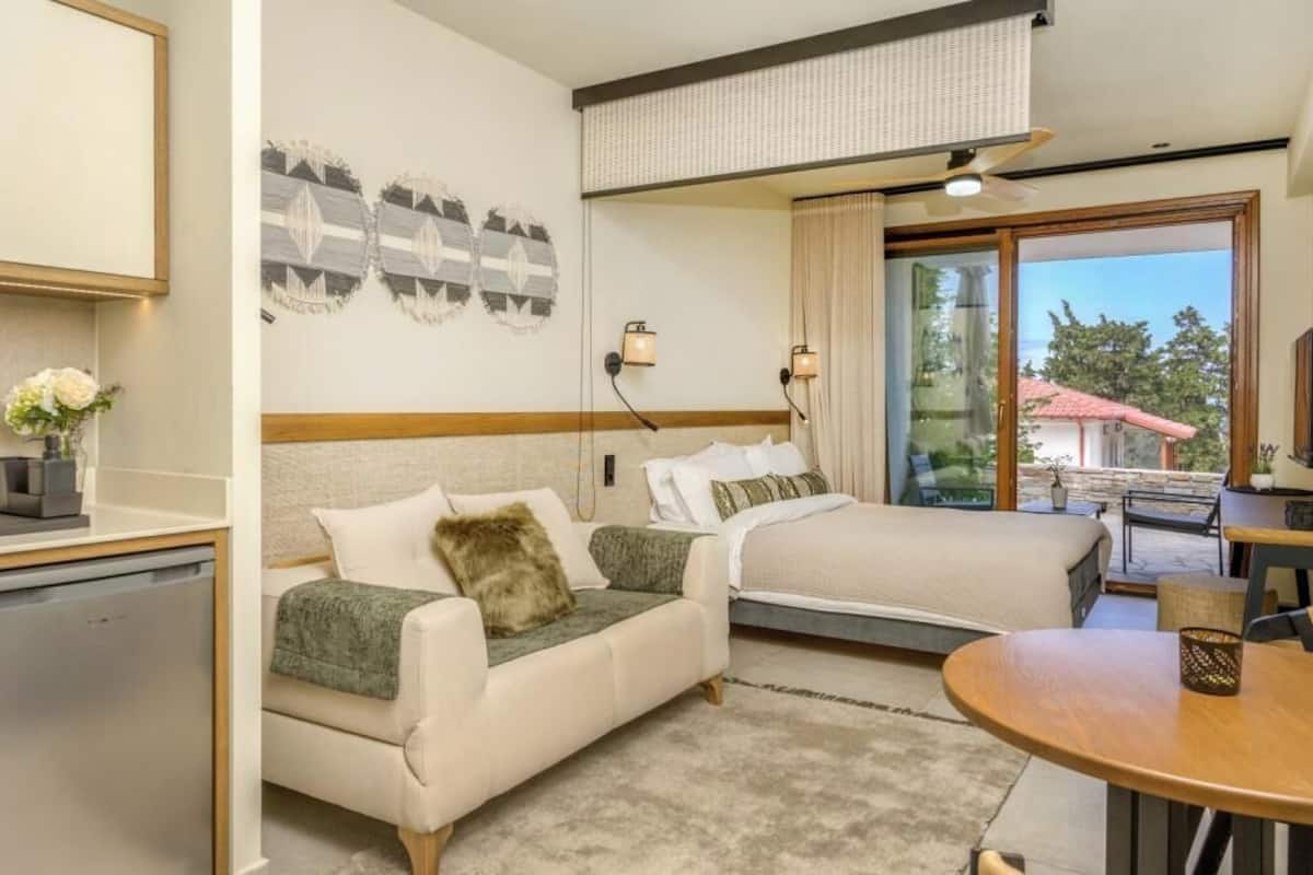 A hotel room with a bed, a sofa, a dining table, and a view of the outdoors.