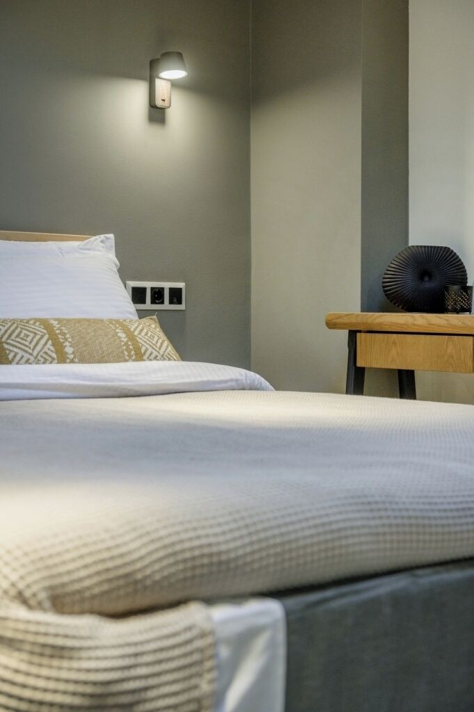 Een netjes opgemaakt bed met een kussen met patroon, een nachtkastje met een lamp en een wandlamp.