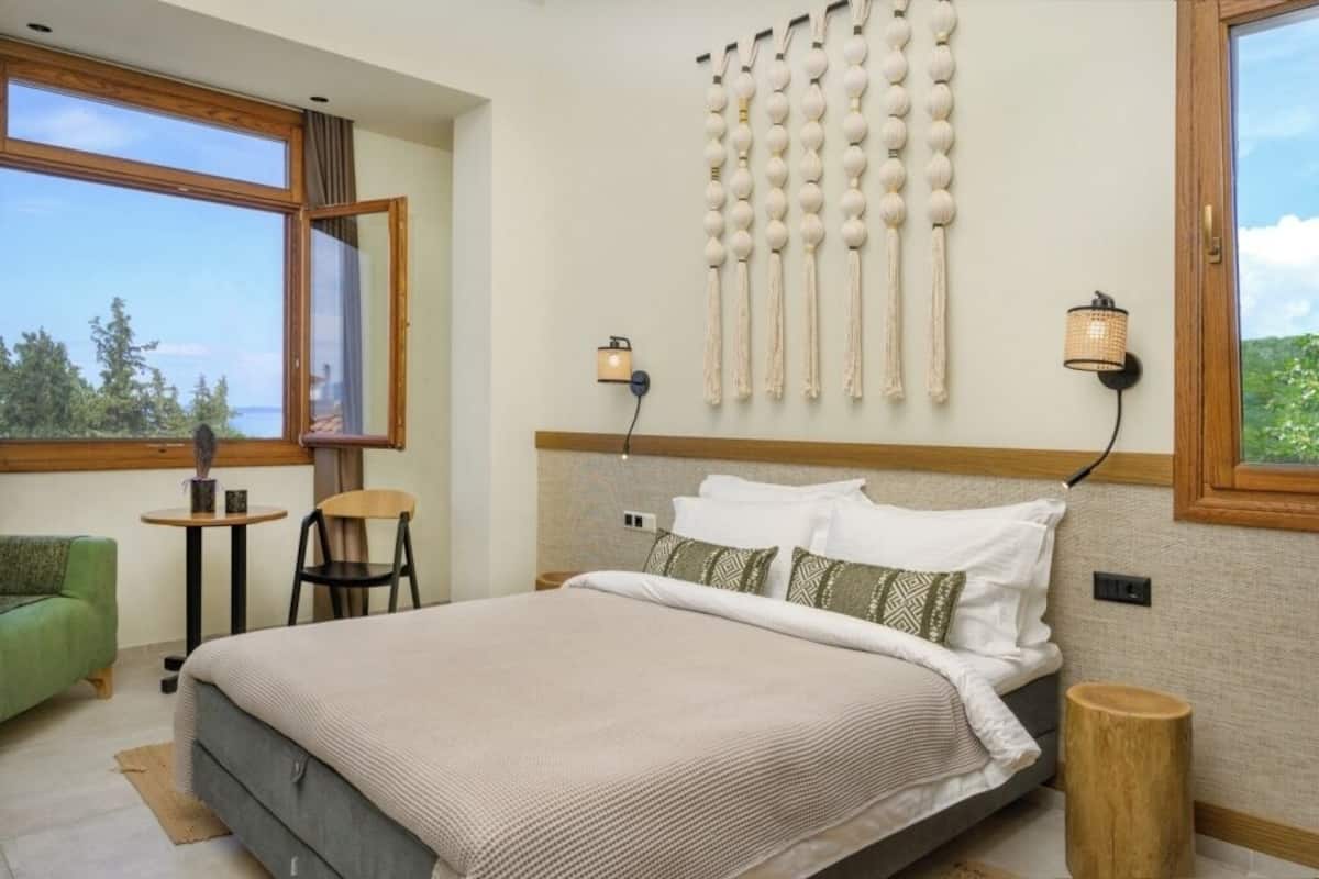 A bedroom with a large bed, a small table, a chair, a decorative wall hanging, and a view through a window.
