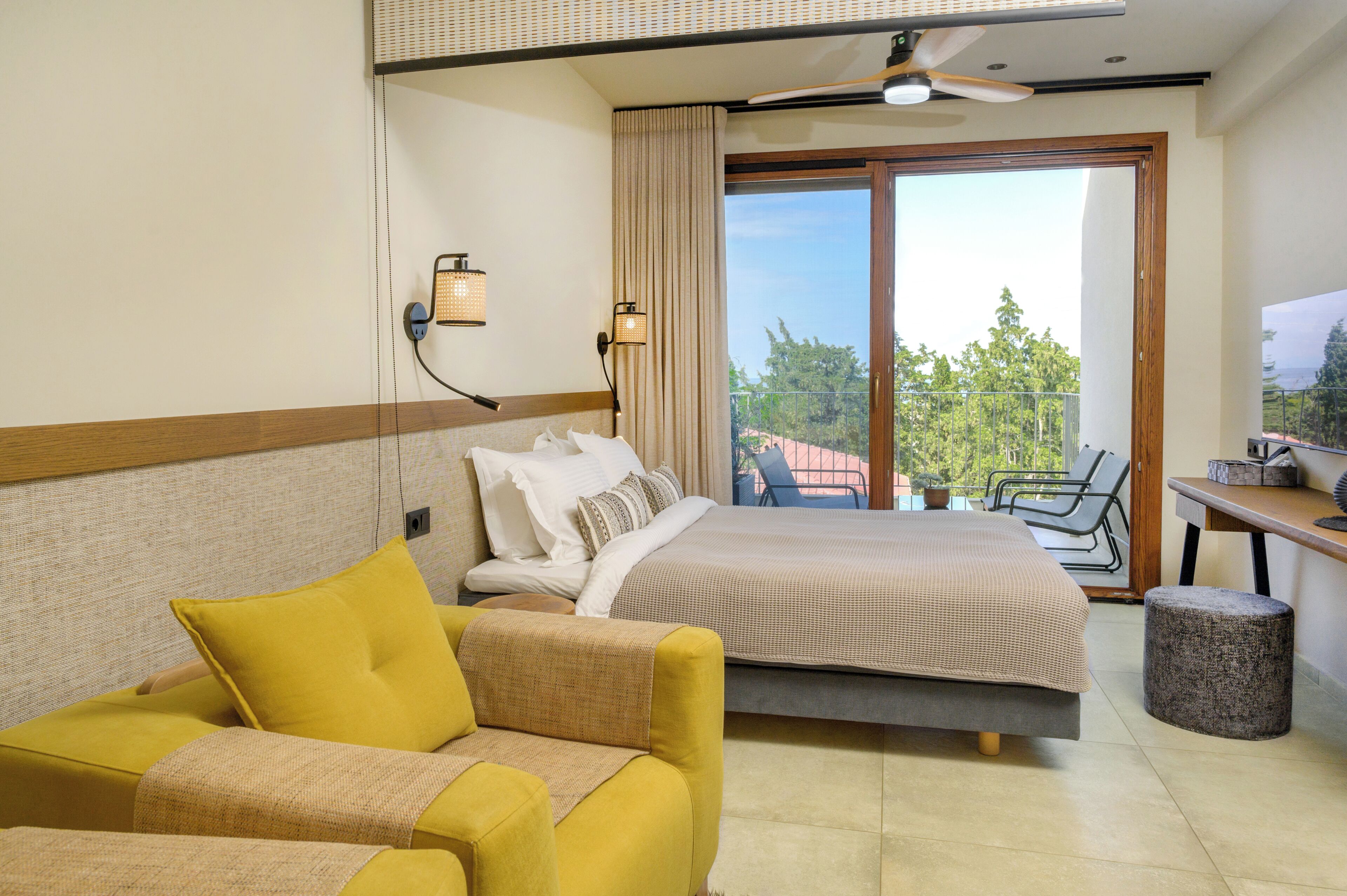 A hotel room with a bed, a sofa, a desk, a chair, a ceiling fan, and a view of the outdoors.